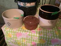 Set of three crocks arranged on table with floral checkered tablecloth. Two beige crocks have visible chips, smaller brown crock with lid is intact.