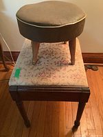 Round brown vinyl upholstered foot stool placed on top of patterned cushioned bench, showing three wooden tapered legs and beige piping trim around the vinyl seat.