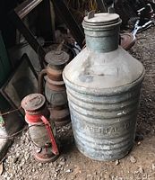 Three vintage lanterns and one large metal Imperial oil can grouped together on a dirt floor showing weathered metal and rust