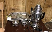 Full lot showing metal coffee percolator, trays, creamer, sugar bowl, and glass condiment servers on a dark wood surface against panel wall