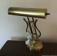 Brass lyre shaped piano lamp with a horizontal cylindrical brass light shade on top and an angel ornament hanging from the lyre frame, placed on a wooden surface.