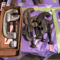 Overview of five pipe cutters laid on table. One RIDGID large pipe cutter in box, four other metal pipe cutters with different sizes and curved handles.