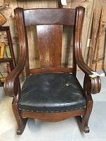 Front view of wooden rocking chair showing wide backrest, curved armrests, black worn seat upholstery.