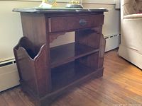 Side angle view of wooden side table showing front drawer, open shelf, and magazine holder attached on side, set against hardwood floor and baseboard heater.
