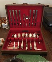 Open view of wooden case lined in red felt containing arranged silverplated flatware pieces with ornate floral handles, showing knives and serving utensils in slots.