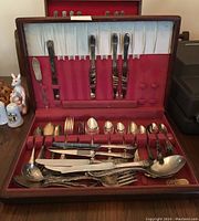 Wooden flatware case lined with red fabric holding approximately 62 mixed pattern silverplated utensils including some serving pieces such as spoons, forks, knives, and specialty utensils.