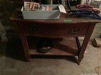 Wooden table with curved front drawer and lower open shelf, situated in basement environment, showing surface wear and scuff marks, with various items on top (not included).