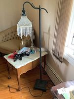 Full view of the vintage floor lamp with frosted glass shade and hanging white bead fringe. The lamp has a bent arm supporting the shade and a square base. It is located next to a wooden side table with decorations.