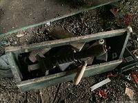 Weathered green metal tool caddy showing its contents from top view with visible large hand tools such as pipe wrench and brace drill inside, surrounded by smaller tools and hardware parts.