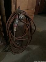 Metal welding tank with coiled rust-colored hose and attached handheld welding torch showing signs of wear.