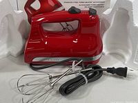 Red KitchenAid hand mixer with power cord and two Turbo Beater stainless steel attachments, placed between white Styrofoam packaging pieces.