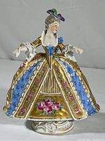 Front and full view of the porcelain figurine, showing the gold and blue dress with floral and striped patterns and lace sleeves.