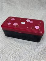 Closed bento box showing red lid with cherry blossom decorations and black base.