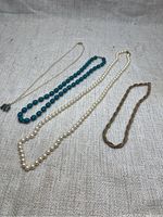 Four necklaces laid out on beige fabric: gold-tone chain with elephant pendant, bold blue bead necklace, creamy faux pearl necklace, and brown braided cord necklace.