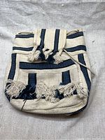 Front view of fabric backpack showing blue and cream horizontal stripes, flap with fringed tassels, front pocket with tassels, drawstring closure.