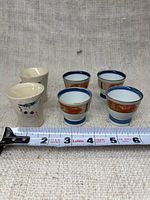 Photo of 6 Japanese sake cups, including 2 white ceramic with black/red design and 4 with blue rims and decorative orange band, sitting on a fabric background with ruler showing size.