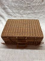 Closed rectangular wicker basket suitcase with handle and latches on front, natural tan color.
