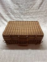 Closed rectangular wicker suitcase basket with front handle and two loop fasteners, viewed from the front on fabric background.