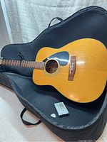 Full view of Yamaha acoustic guitar lying in black soft case with digital guitar tuner and white guitar pick visible