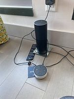 Photo showing large black cylindrical Amazon Echo and smaller white Amazon Echo Dot on floor with power cords and an Echo manual.