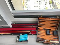 Two pool cues in black cases alongside an open backgammon set and a vintage domino box on the ground.