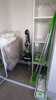Wide view of corner showing Bissell Bolt vacuum, two dry mops, pack of dry mop pads, white shelf unit, and curtain rod.