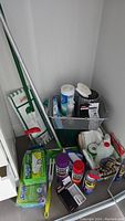 Full view of corner with multiple cleaning supplies including Swiffer mop/pads, cleaning sprays, gloves, and brushes.