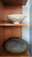 Shelf view of white ceramic abstract swirl bowl on upper shelf and large mottled green/brown bowl below
