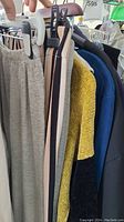 Photo showing a rack with multiple hanging garments, including a yellow knit sweater, navy blue knit, black jacket, and striped trousers.