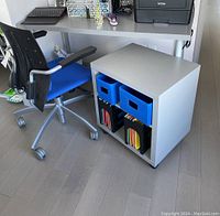 Desk with gray metal legs and smooth surface next to a rolling file cabinet with two blue bins and hanging files.