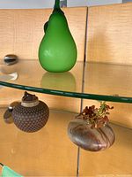 Glass pear, small glass vase with dried flowers, and patterned clay pot displayed together on glass shelf.