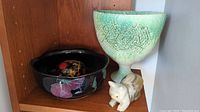 Photo showing the green footed bowl, black floral bowl with decorative egg inside, and small cat carving figurine on a shelf.