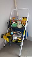 Full lot of home repair items arranged on and around a white metal frame step stool/cart including tapes, finishing wax, staple gun, and a wooden broom or mop handle.