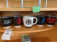 Front view of 5 Indigenous art coffee mugs on shelf, 4 black mugs and 1 white mug with distinct Indigenous art designs
