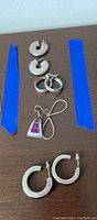 Five pairs of ladies earrings arranged on table with blue tape as reference, showing different styles and finishes.