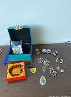 Overview of jewelry lot showing green jewelry box, wooden bead bracelet, earrings, pendant, guitar pick, and coins.
