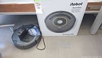 Robot vacuum and charging dock positioned in front of original box; spare filter pack visible to side