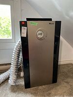 Full view of portable air conditioner with attached flexible exhaust hose and power cord on carpeted floor near window.