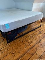 View of bed frame with mattress showing black metal frame and white mattress top with visible blue branding on mattress side.