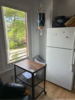 Rolling cart with wood-look top surface and two mesh shelves, positioned next to a refrigerator and under a window.