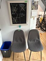View of two gray upholstered dining or side chairs placed side by side beneath a framed chalkboard affixed to the wall. The chalkboard has lists written in white chalk and a small greenish-blue mini pail attached under the frame.