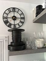 Black metal wall clock with Roman numerals and gold hands mounted on wall above shelves with stacked black stoneware plates and bowls, assorted clear glassware and white and green bowls.
