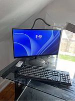 Acer 21 inch monitor lit on screen with screen showing 9:49 time, black round base, black wired Logitech keyboard in front on glass desk with white adjustable desk lamp.