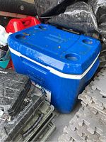 Blue Coleman cooler with lid, visible carrying handle and cup holders, placed on gray foam floor mats.