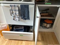 Kitchen stove open drawer with baking sheet and loaf pan inside, open cabinet with Crock Pot visible
