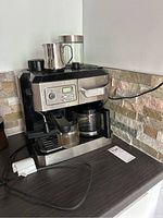 DeLonghi coffee and espresso machine placed on countertop with glass coffee pot, metal milk frothing pitcher, and glass canister on top.