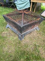 Front angle photo of black metal square fire pit with mesh lid showing patterned sides and legs. Exhibits rust and wear, placed on grass.
