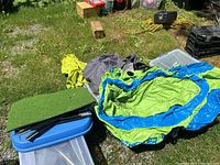 Outdoor image showing green golf tee-off mat, disassembled black poles for target net, Pathfinder inflatable boat material in green and blue, all placed on large plastic tote with other items around.