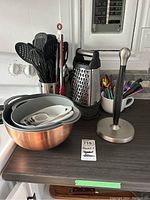 Full lot on kitchen counter showing metal mixing bowl, nested plastic bowls and measuring cups, metal box grater, paper towel holder, utensil holder with plastic utensils, cafe au lait mug with cutlery, metal tongs.