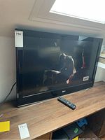 Front view of the LG 37 inch television switched on showing a program on the screen, with remote control placed on the wooden surface in front.